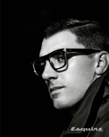 Close-up black and white portrait of a man wearing thick dark eyeglasses and a high-collared jacket against a dark background