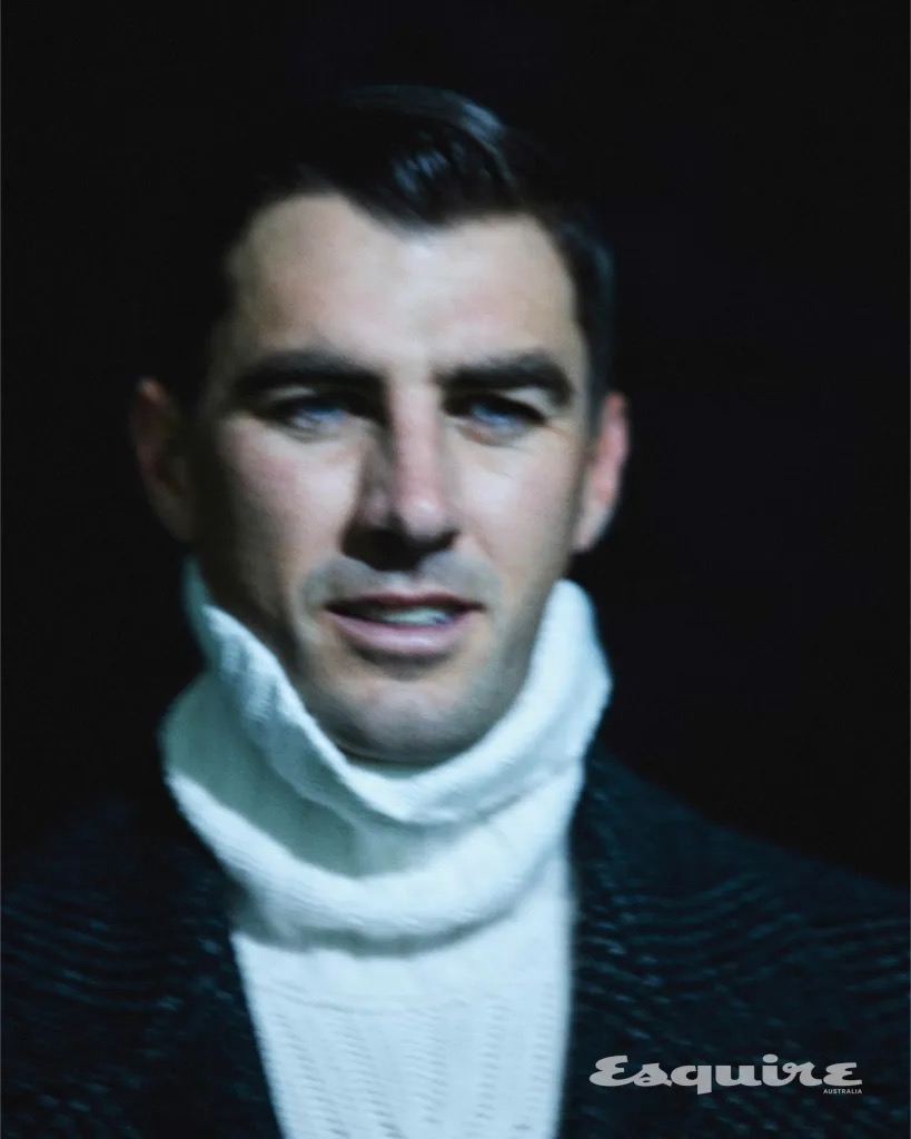 Portrait of a man with short dark hair, wearing a white turtleneck sweater and a dark coat against a dark background.