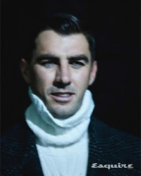 Portrait of Pat Cummins wearing a white turtleneck sweater and dark patterned jacket against a dark background.