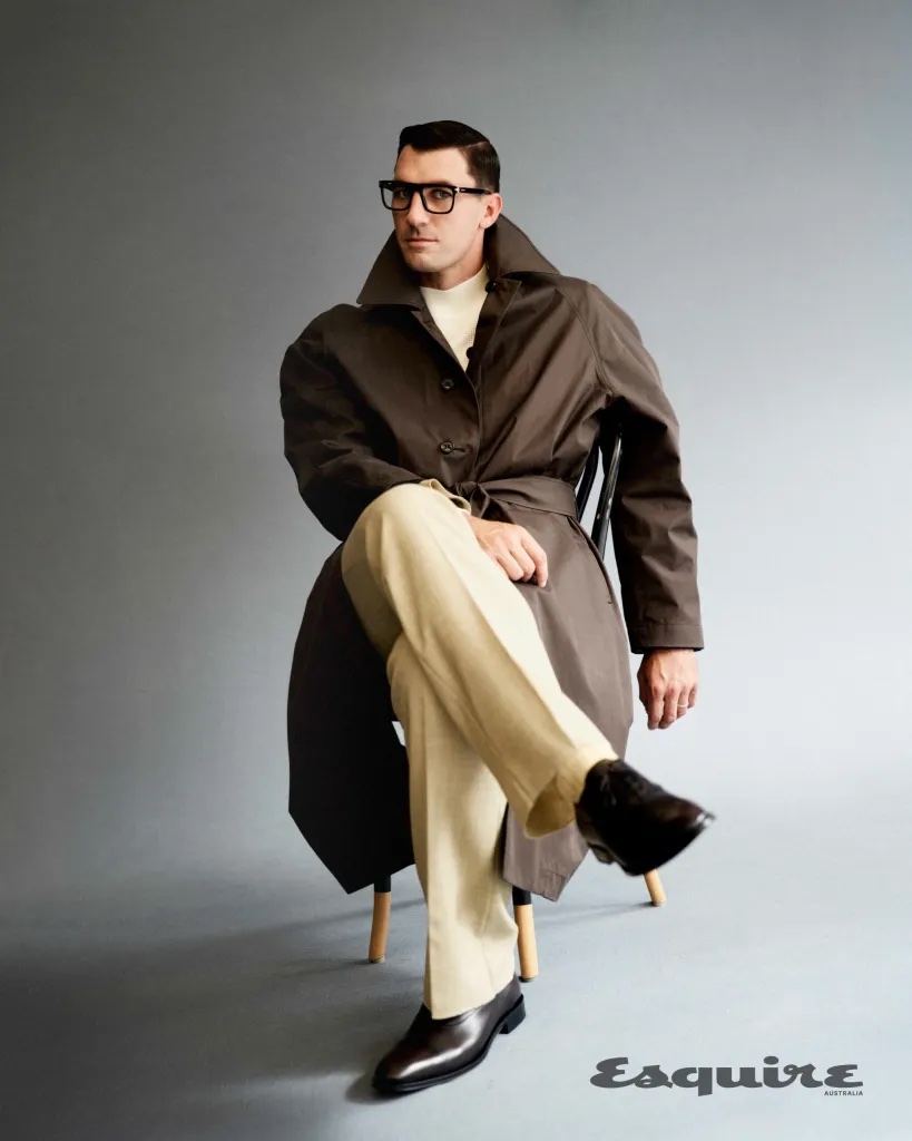 An adult male wearing glasses and a trench coat is sitting on a wooden chair in a photo studio with a solid grey background.