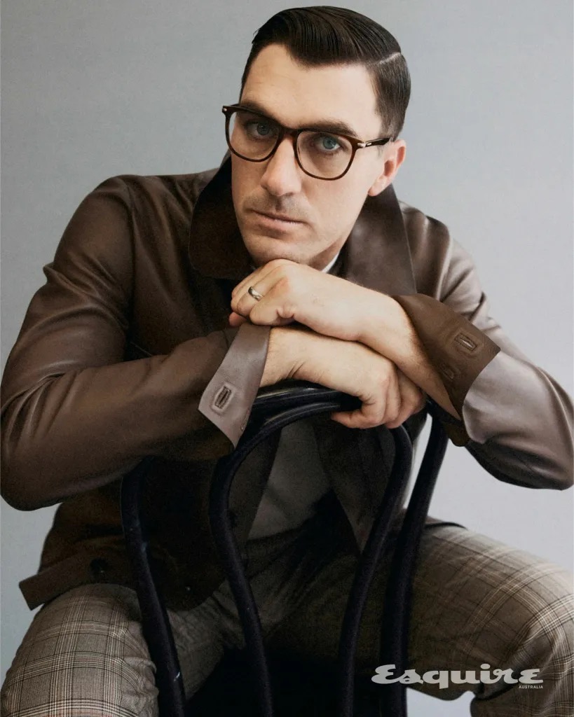 Man wearing glasses and a brown leather jacket resting his arms on the back of a chair against a grey studio wall.