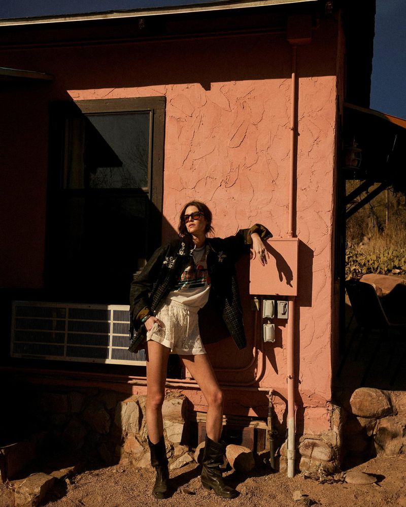 Adult female in a jacket and shorts poses against a textured wall of an outdoor building in sunlight with visible desert plants.