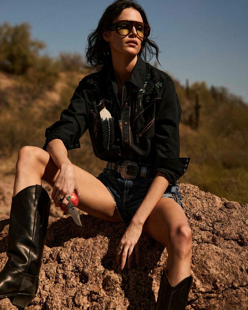Woman in cowboy boots sitting on a rock in desert landscape wearing a black western shirt, jean shorts, and sunglasses.