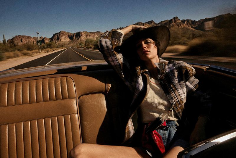 A woman in a hat and flannel poses in a convertible car on a desert highway with mountains in the background, bathed in sunlight.