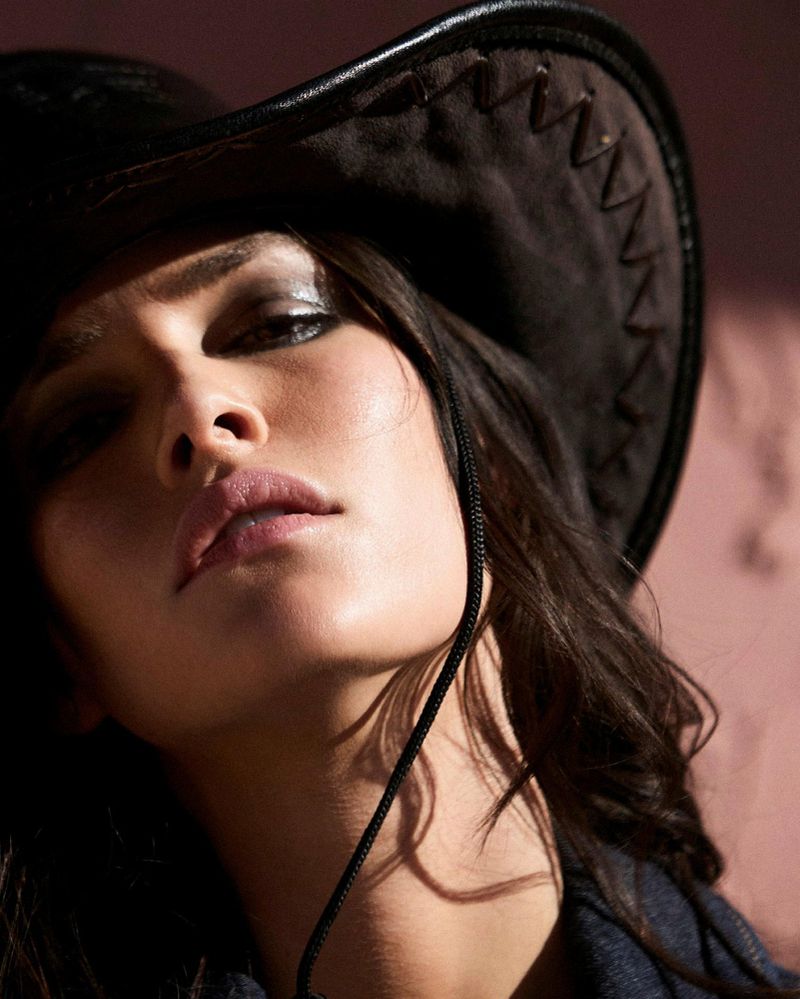 Close-up portrait of a brunette adult woman wearing a dark cowboy hat in a studio, with dramatic lighting highlighting her makeup.