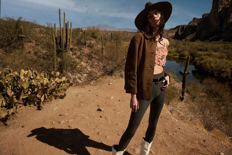 A young woman poses in a desert landscape wearing a hat, jacket, leggings, a floral crop top, and boots on a sandy path.