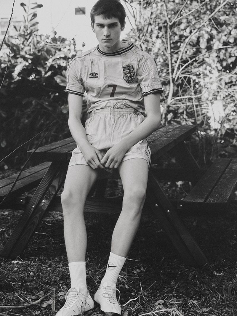 A young adult male sits on a wooden picnic table outside, wearing a vintage soccer jersey, shorts, socks, and tennis shoes. The background features green foliage.