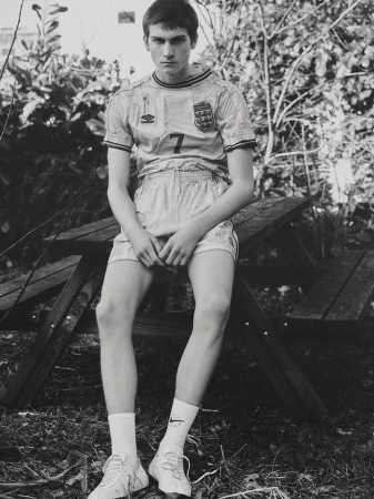 A young adult male sits on a wooden picnic table outside, wearing a vintage soccer jersey, shorts, socks, and tennis shoes. The background features green foliage.