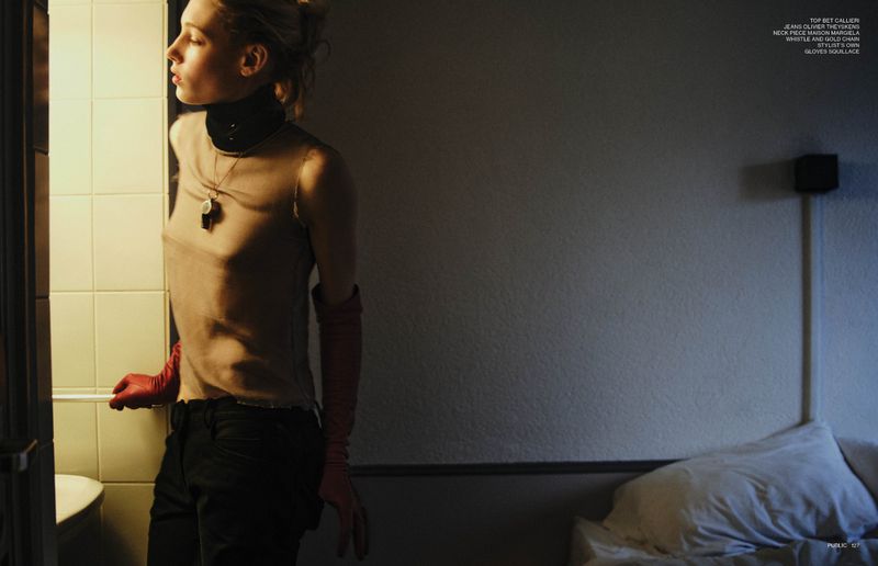 Portrait of a blonde woman in a tan top, black pants and red gloves, standing indoors with fair skin and looking out of frame.