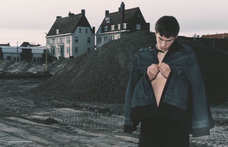 A young man poses outdoors in a denim jacket and black pants, standing on a dirt mound in front of buildings under an overcast sky.