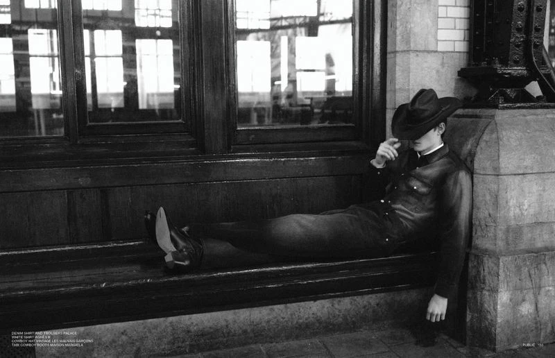 Model wearing a cowboy hat and dark denim resting horizontally on a wooden bench inside a transit station