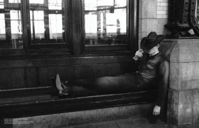 Model wearing a cowboy hat and dark denim resting horizontally on a wooden bench inside a transit station
