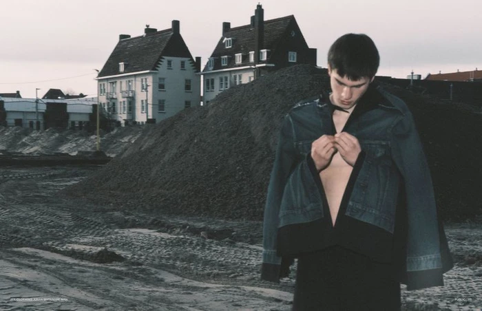 Young man buttoning a dark jacket outdoors in front of a dirt mound and residential houses under an overcast sky.