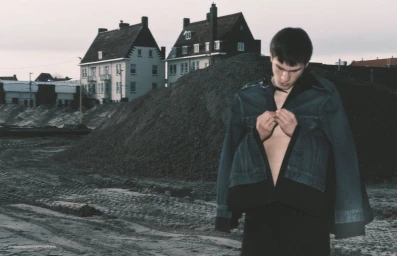 Young man buttoning a dark jacket outdoors in front of a dirt mound and residential houses under an overcast sky.