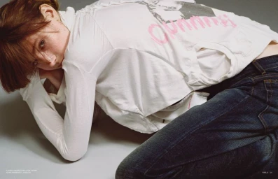 Model in white long-sleeve graphic top and dark denim jeans posing on a grey studio floor for PUBLIC Editions Dutch Tailgate.