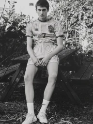 Young man in football jersey and shorts seated on a wooden picnic table outdoors in a monochrome editorial portrait.