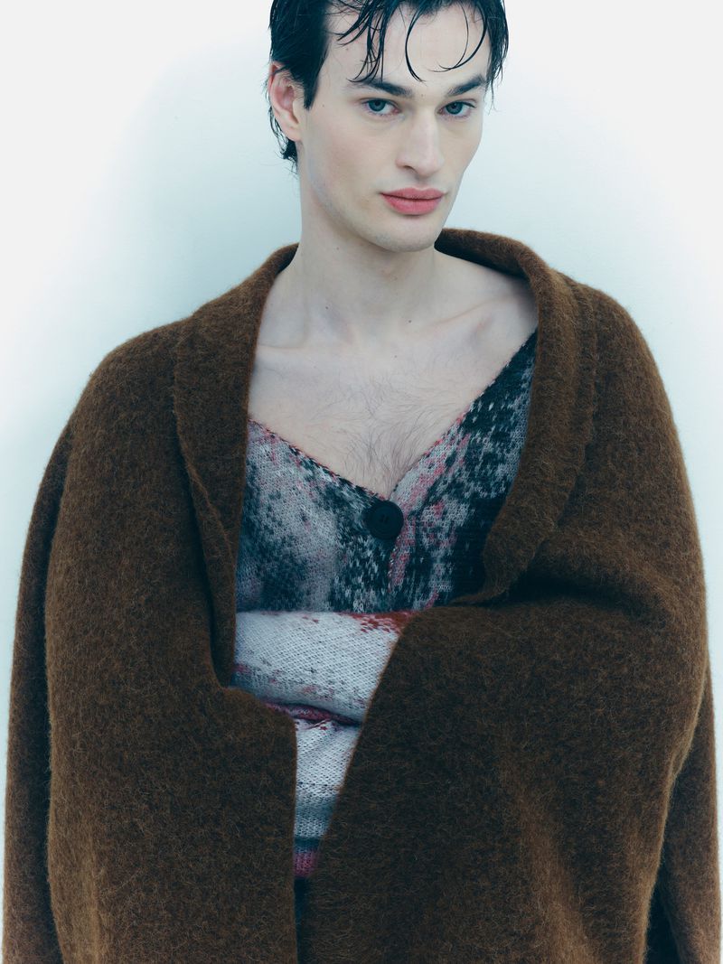 Portrait of a young adult male in an open brown mohair cardigan over a printed top, standing in front of a plain white wall.