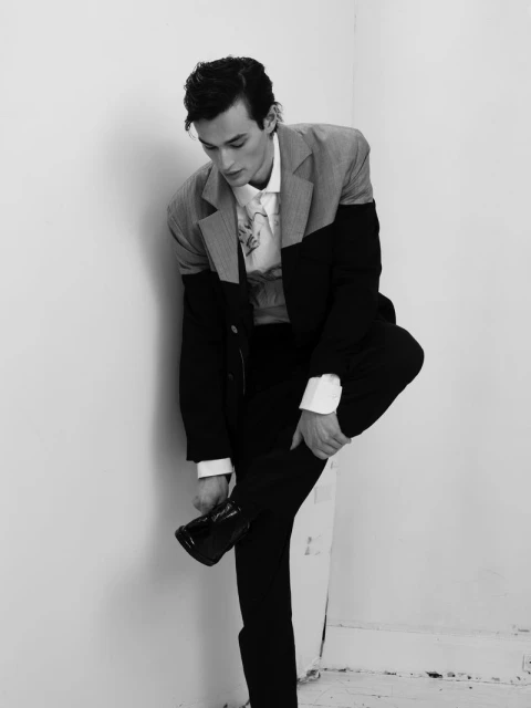 Monochrome portrait of a young adult man wearing a black and grey suit, putting on a shiny black dress shoe in a white corner.