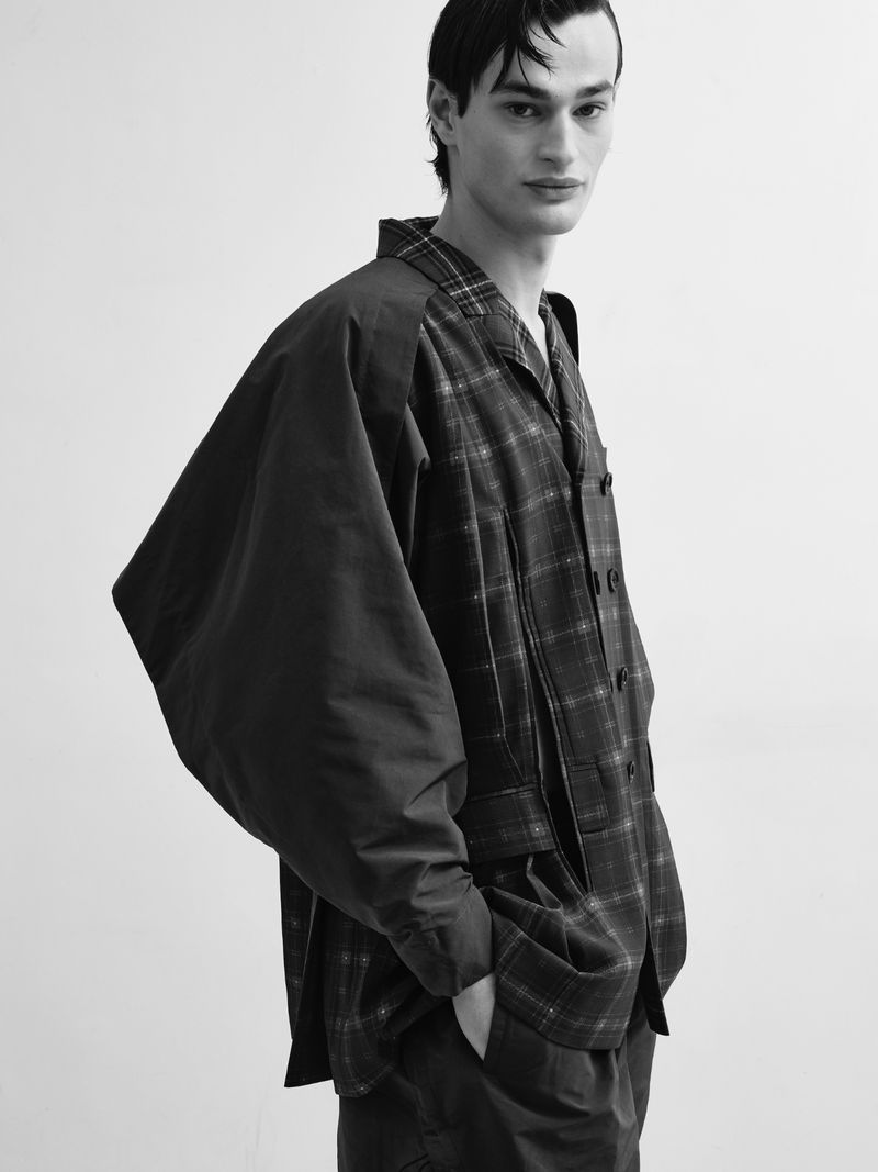 Monochrome portrait of a male model in a plaid jacket and loose pants against a white backdrop, captured in a studio setting.