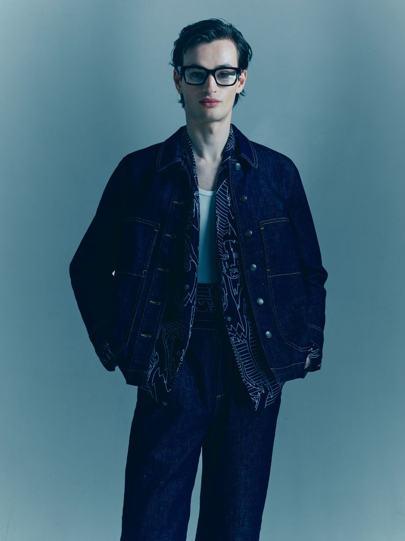 A slender adult male model poses in a photo studio, wearing a denim jacket, matching pants, and black rectangular glasses against a neutral backdrop.