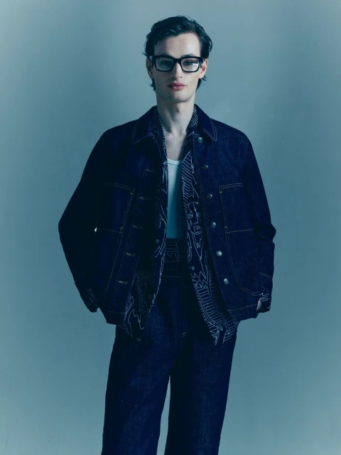 A slender adult male model poses in a photo studio, wearing a denim jacket, matching pants, and black rectangular glasses against a neutral backdrop.