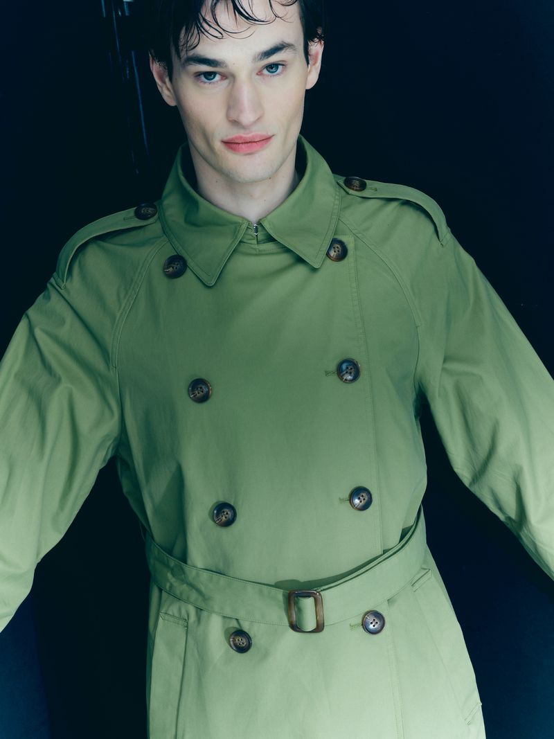 Portrait of a fashionable adult male wearing a green trench coat against a dark backdrop in a studio setting with cinematic lighting.