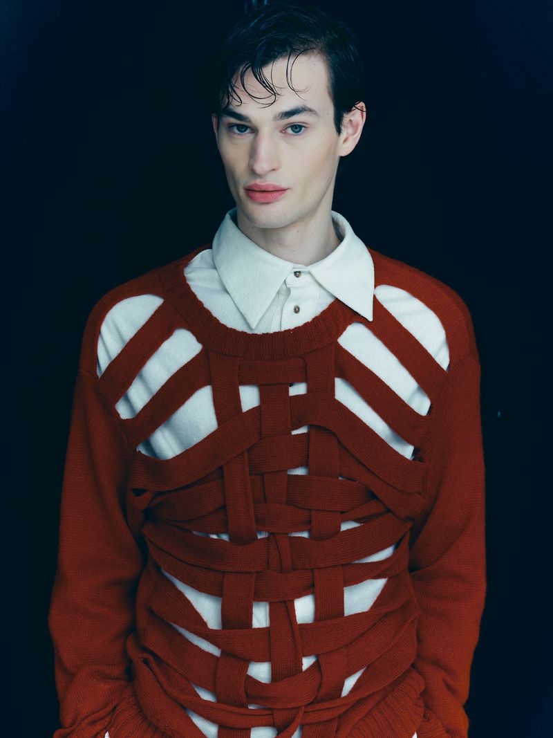 Portrait of a young adult man with slicked-back dark hair wearing a cream-colored collared shirt and deconstructed knit sweater, in studio.