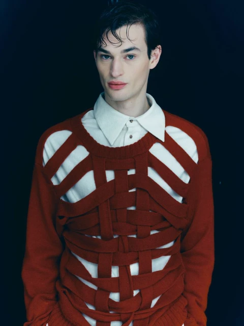 Portrait of a young adult man with slicked-back dark hair wearing a cream-colored collared shirt and deconstructed knit sweater, in studio.