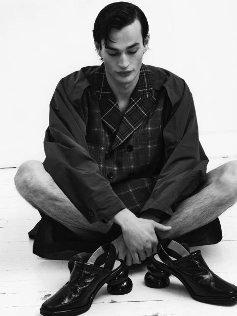 Monochrome shot of a man seated with crossed legs, gazing down at a pair of high-heeled shoes on a white studio floor.