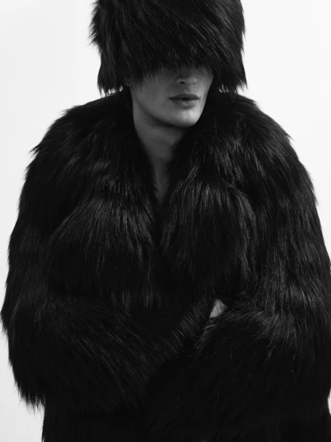 Black and white portrait of a slender adult male wearing a large, dark fur coat and a furry hat that obscures his eyes.