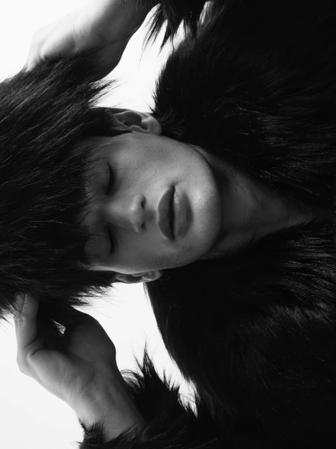 Monochrome portrait of a young adult male with closed eyes, hands on his head, wearing a faux fur coat against a bright white background.