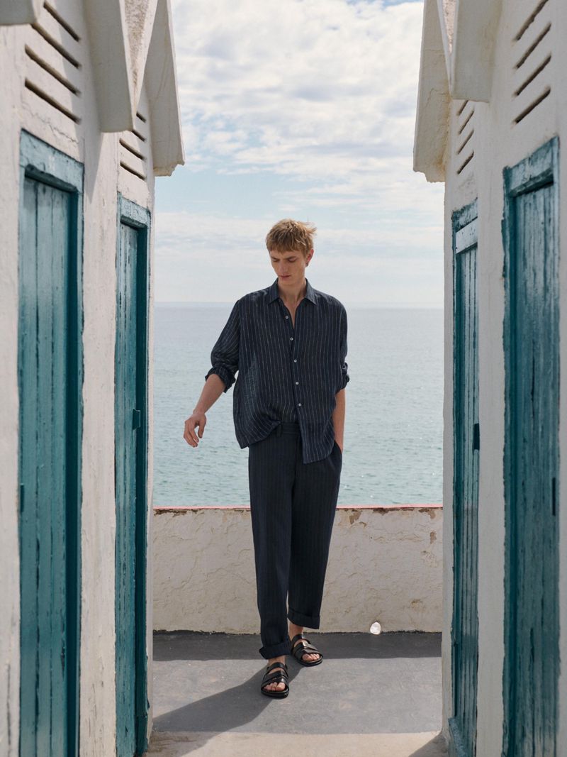 A blond male model walks toward the camera in a relaxed pose near beachside huts, wearing a striped shirt and pants with leather sandals.