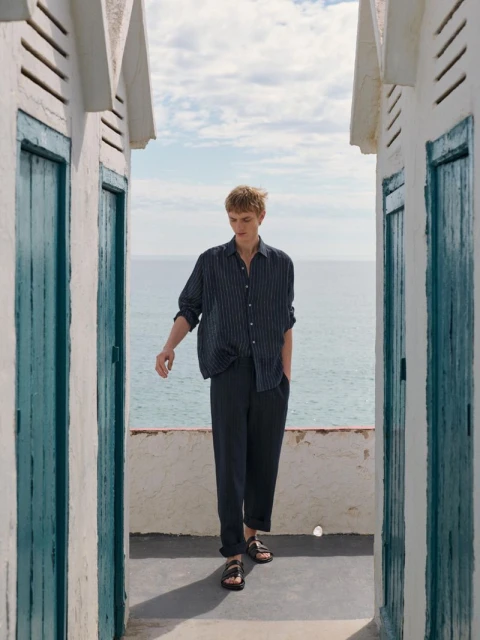 A blond male model walks toward the camera in a relaxed pose near beachside huts, wearing a striped shirt and pants with leather sandals.