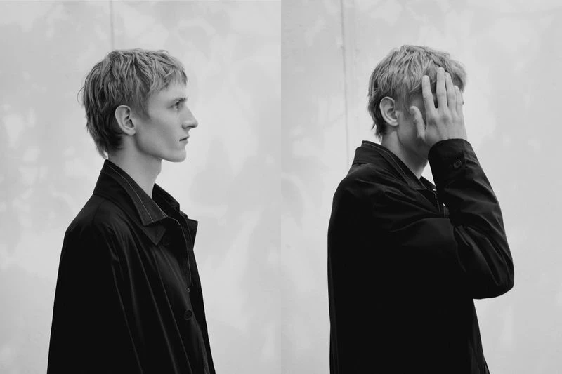 Monochrome portrait of a slender adult man with short blond hair wearing a dark collared jacket against a neutral background, split view.