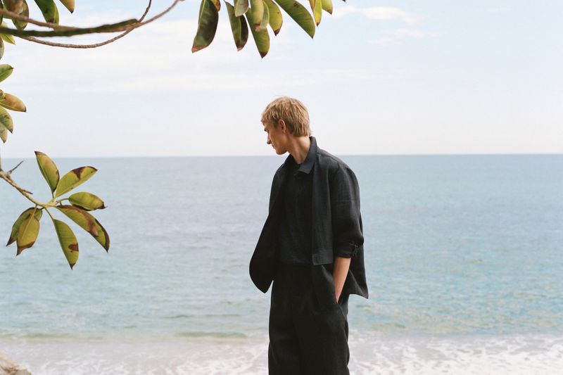 Young adult male model in black linen suit stands near the sea, framed by tree branches and leaves, looking away on a bright day.