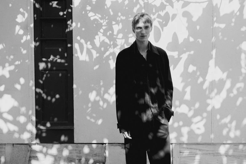 Monochrome portrait of a slender, light-skinned adult male in a dark casual outfit standing in front of a wall with leafy shadows.