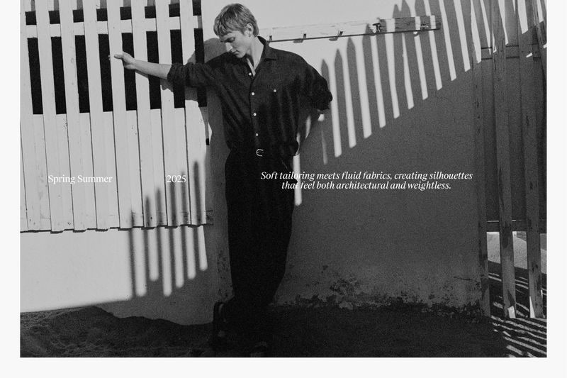 A young man in black shirt and pants leans against a white fence on a beach, with shadows casting striking lines, promoting fashion.