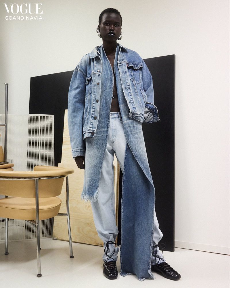 Full-length shot of a Black woman in a light-wash denim outfit, statement earrings, and black shoes in a photo studio setting.