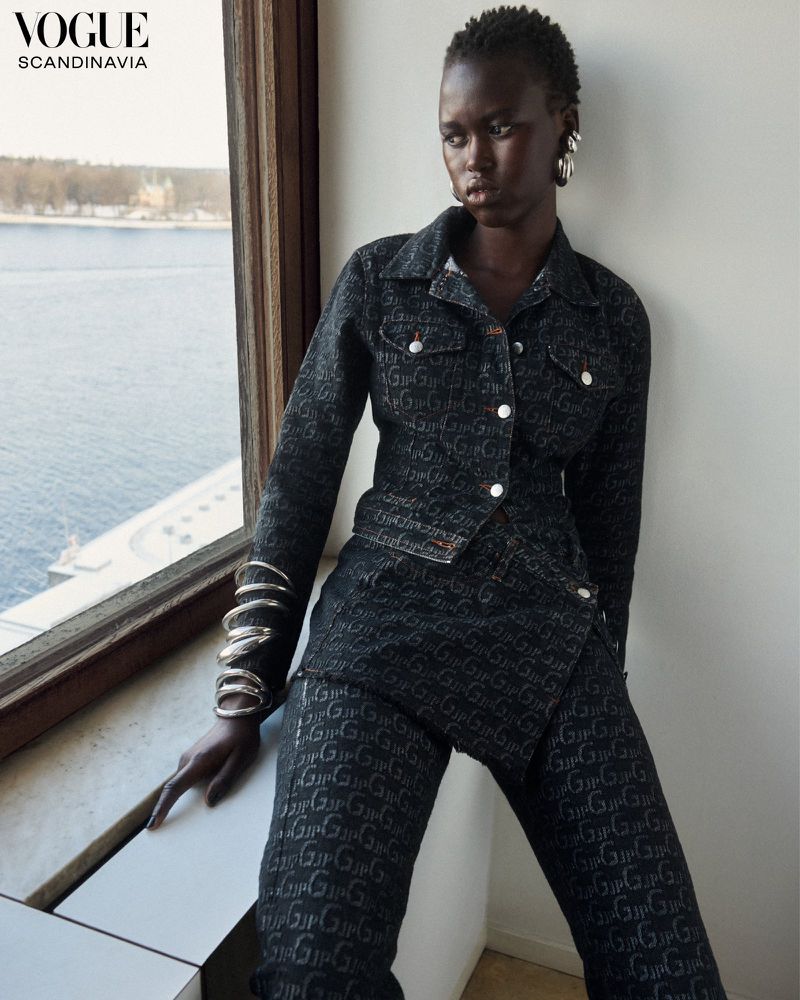 Editorial fashion shot of a dark-skinned woman in patterned jacket and pants, posing by a bright window with a city view.