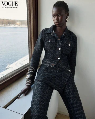 Editorial fashion shot of a dark-skinned woman in patterned jacket and pants, posing by a bright window with a city view.