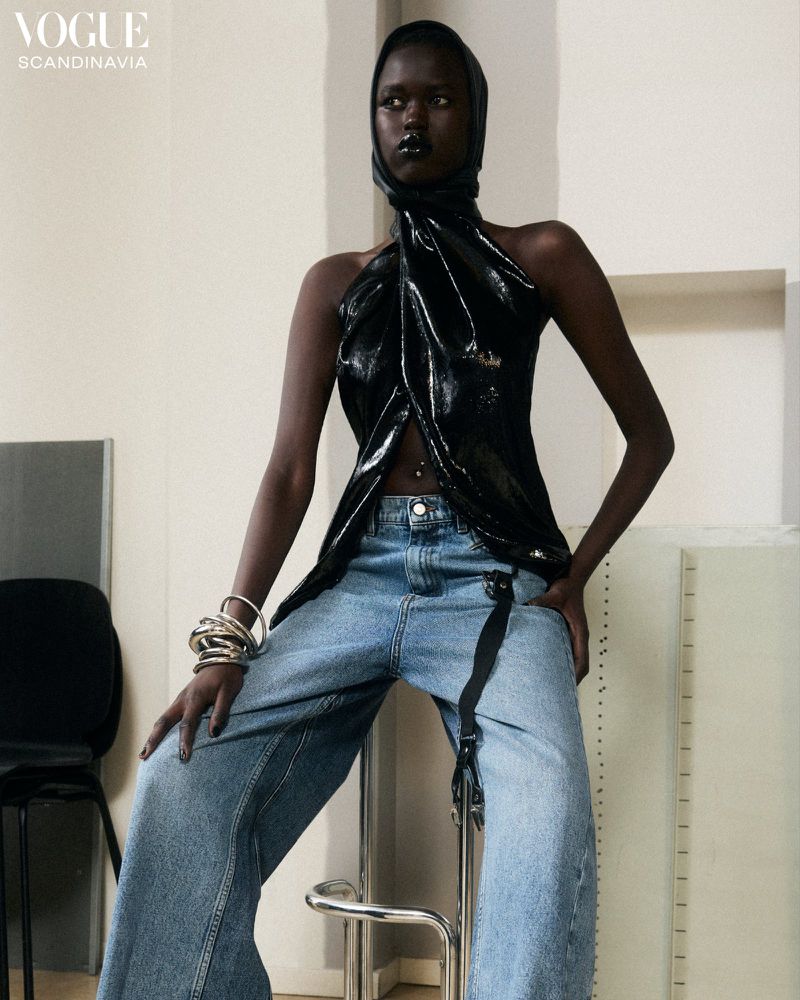 A fashion shot of a black female model wearing a shiny black top and baggy jeans while sitting on a stool in a studio environment.
