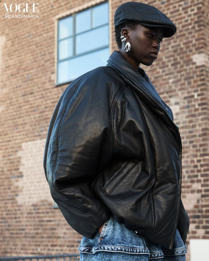 Fashion portrait of a Black adult female wearing an oversized leather jacket, newsboy cap and dangling earrings, set against a brick building.