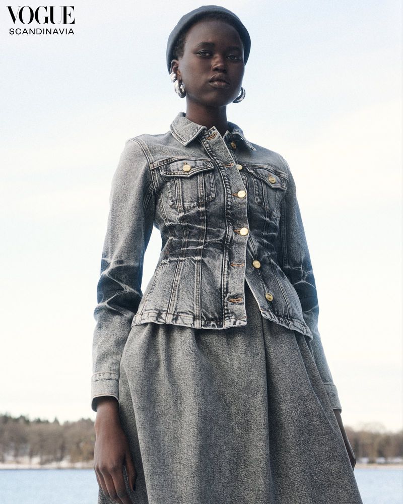 Woman in gray denim jacket and dress poses outdoors, wearing a black beret and hoop earrings in a fashion editorial shot.