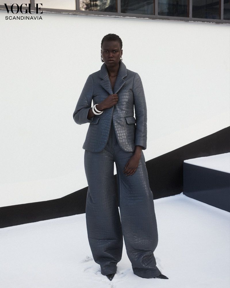 Woman in an all-grey outfit posing against a white backdrop, fashion editorial shot. She's wearing a textured jacket and wide-legged trousers.