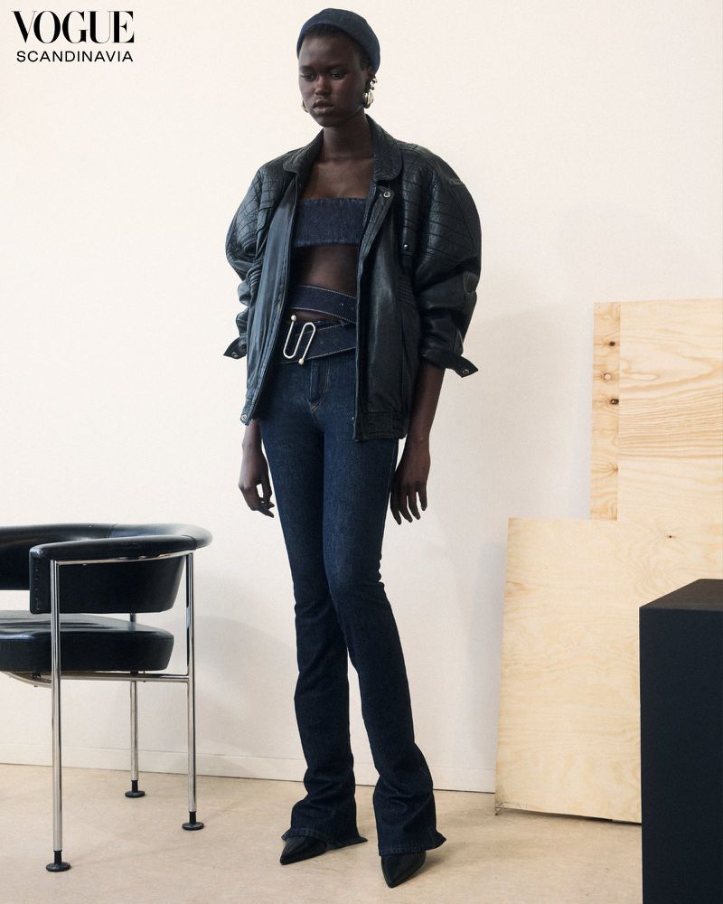 An adult woman poses in a fashion editorial shot wearing a dark jacket, denim top, and flared jeans against a neutral wall.