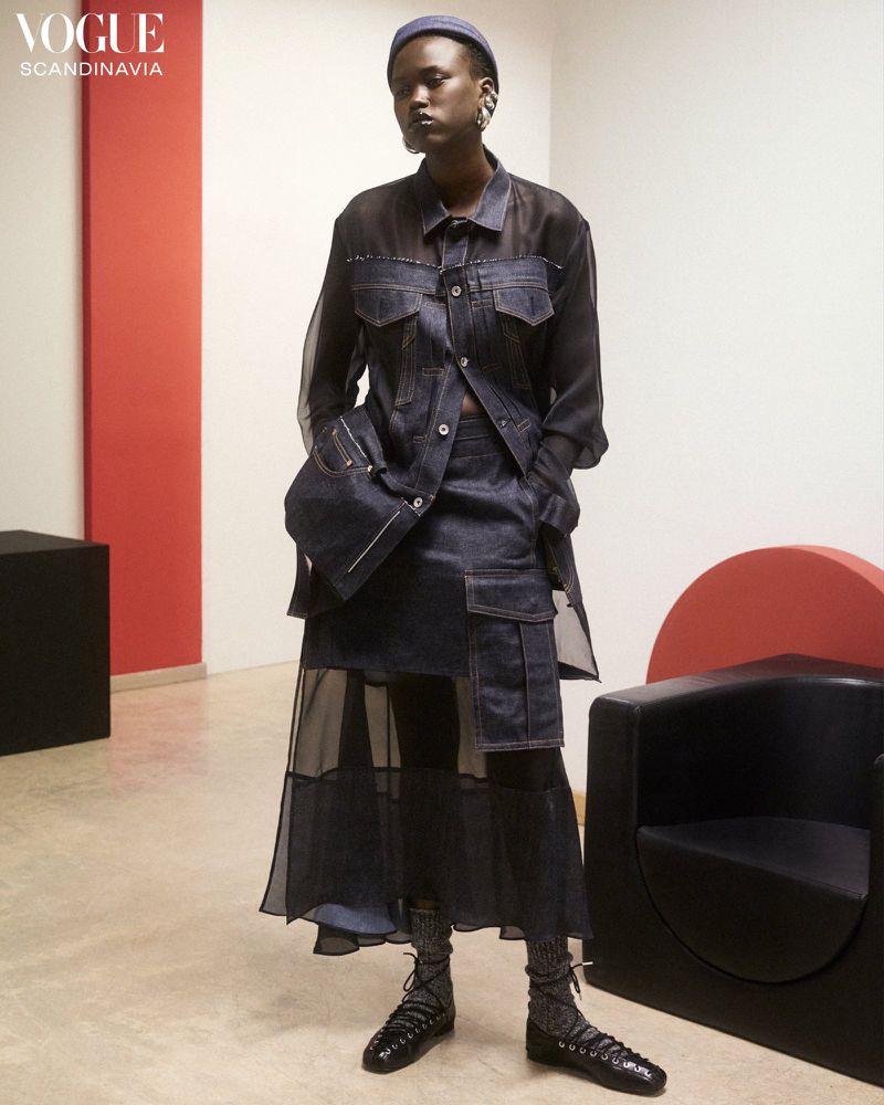 Fashion editorial shot of a Black female model wearing a layered blue denim and sheer ensemble, posing in an indoor studio setting.