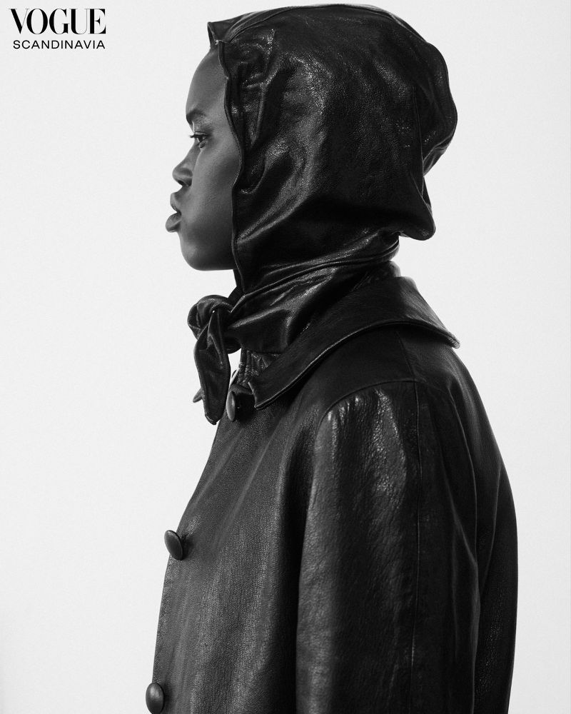 Profile shot of a woman with dark skin wearing a black leather coat and hood against a bright white background in a studio setting.