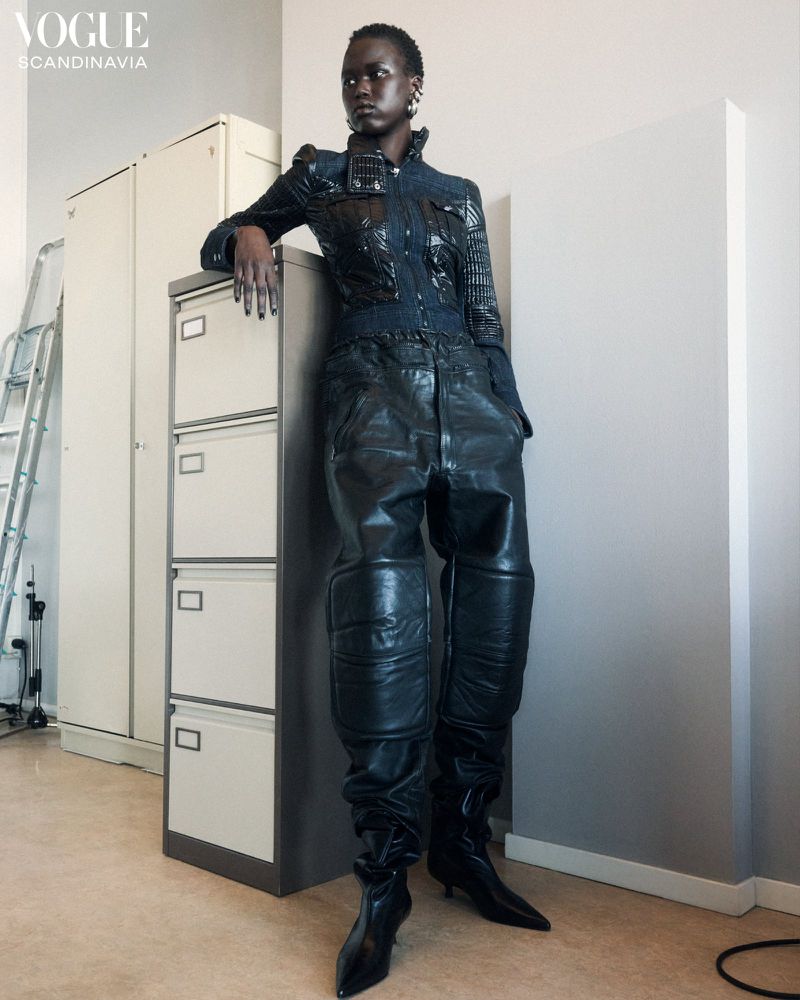 A striking editorial fashion shot of a model in a dark jacket and pants leaning against a filing cabinet in a minimalist indoor setting.