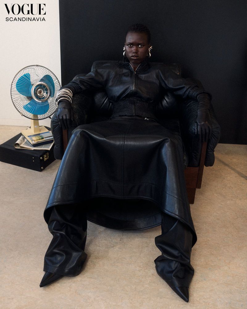 Dark-skinned adult woman in a leather coat and skirt sits in a chair in front of a black and white wall and a vintage fan.