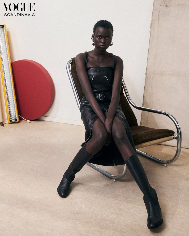 An adult woman poses in a black leather dress and boots, seated on a tubular chrome chair in a minimalist studio setting for Vogue Scandinavia.